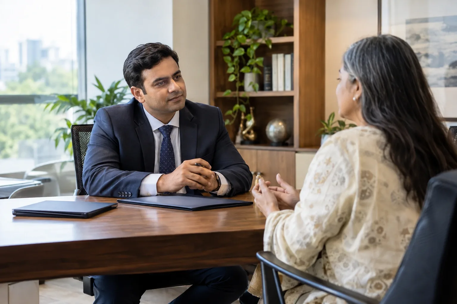 An Indian financial adviser listening to an adult client at a tidy office desk, blank folder and tablet face down, natural light, measured trustworthy mood.